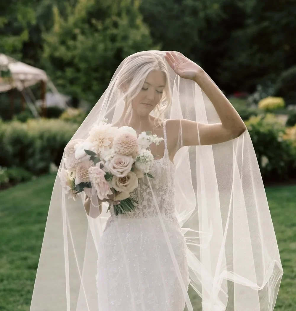 A bride in a white wedding dress holding a bouquet of pink and white flowers, standing outdoors with a veil over her face and surroundings of green trees.