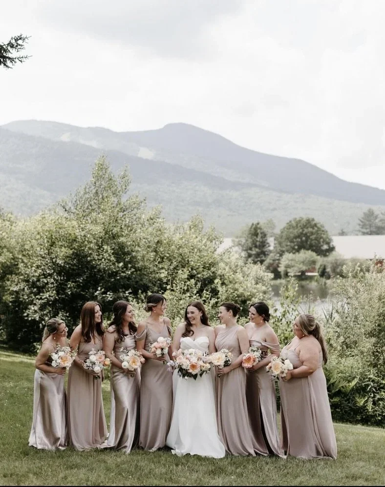 A bride in a white wedding dress surrounded by seven bridesmaids in matching mauve dresses, outdoors with green trees, grass, and mountains in the background.