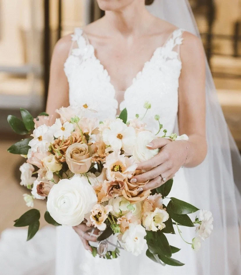 A bride holding a bouquet of pastel-colored flowers including roses, ranunculus, and other blooms in soft peach, white, and cream shades, with green leaves, wearing a white lace wedding dress with floral straps.