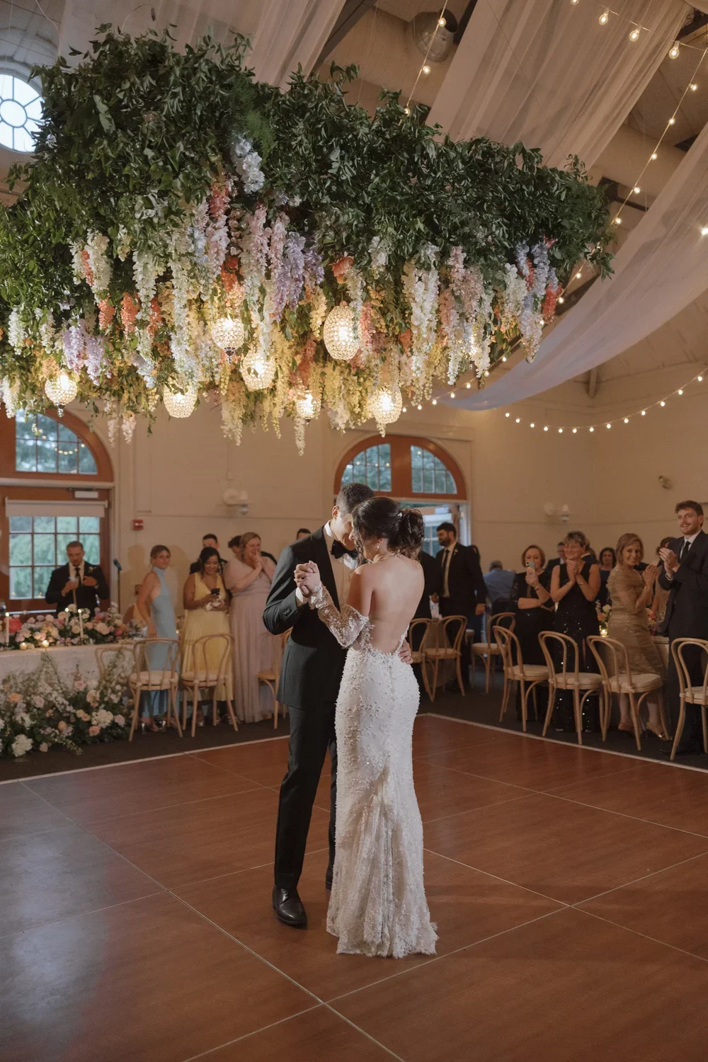 A bride and groom share a dance during their wedding reception, surrounded by guests clapping and smiling, beneath a large hanging floral arrangement with lights.