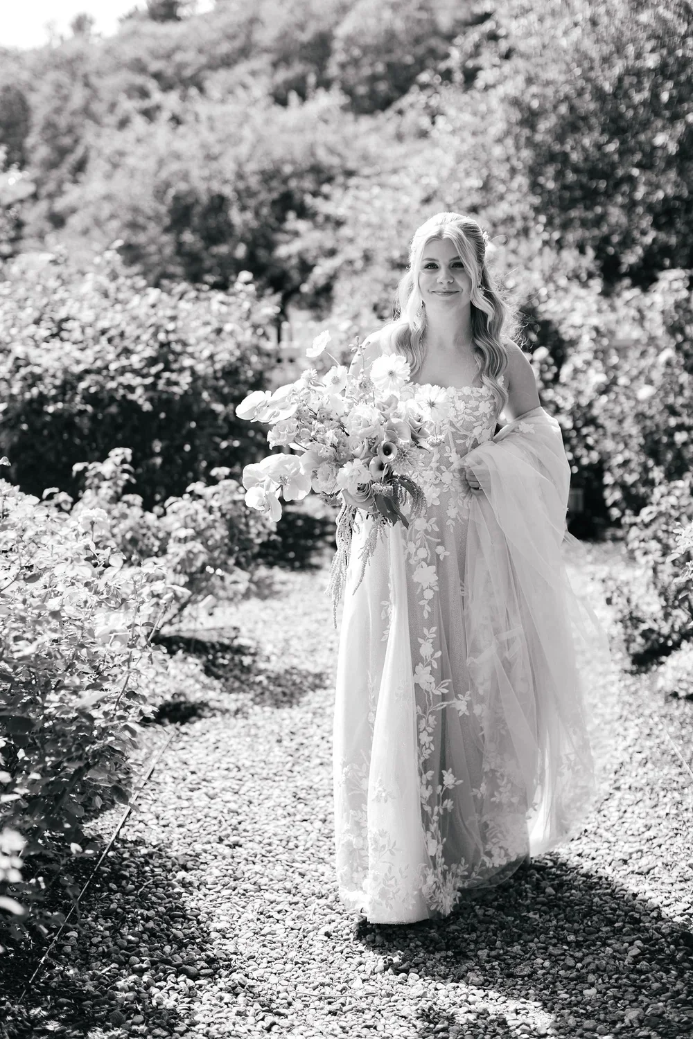 A bride walking through a garden, holding a bouquet of flowers, wearing a lace dress and a tulle veil, surrounded by lush foliage.