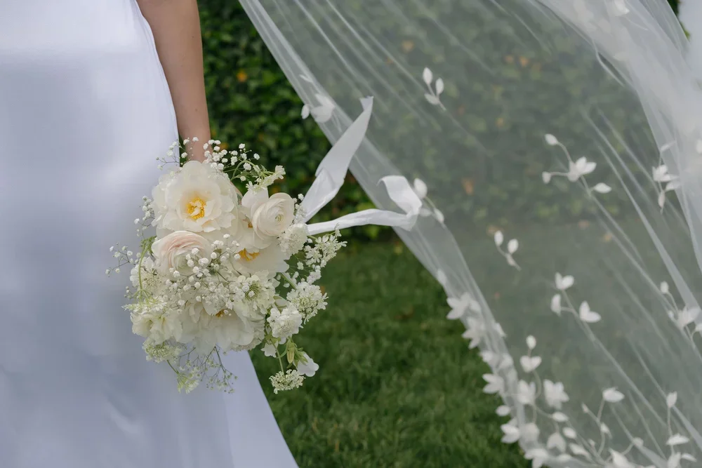 A bride holding a white bouquet with roses, peonies, and baby's breath, standing outdoors with a veil decorated with small white floral patterns flowing nearby.