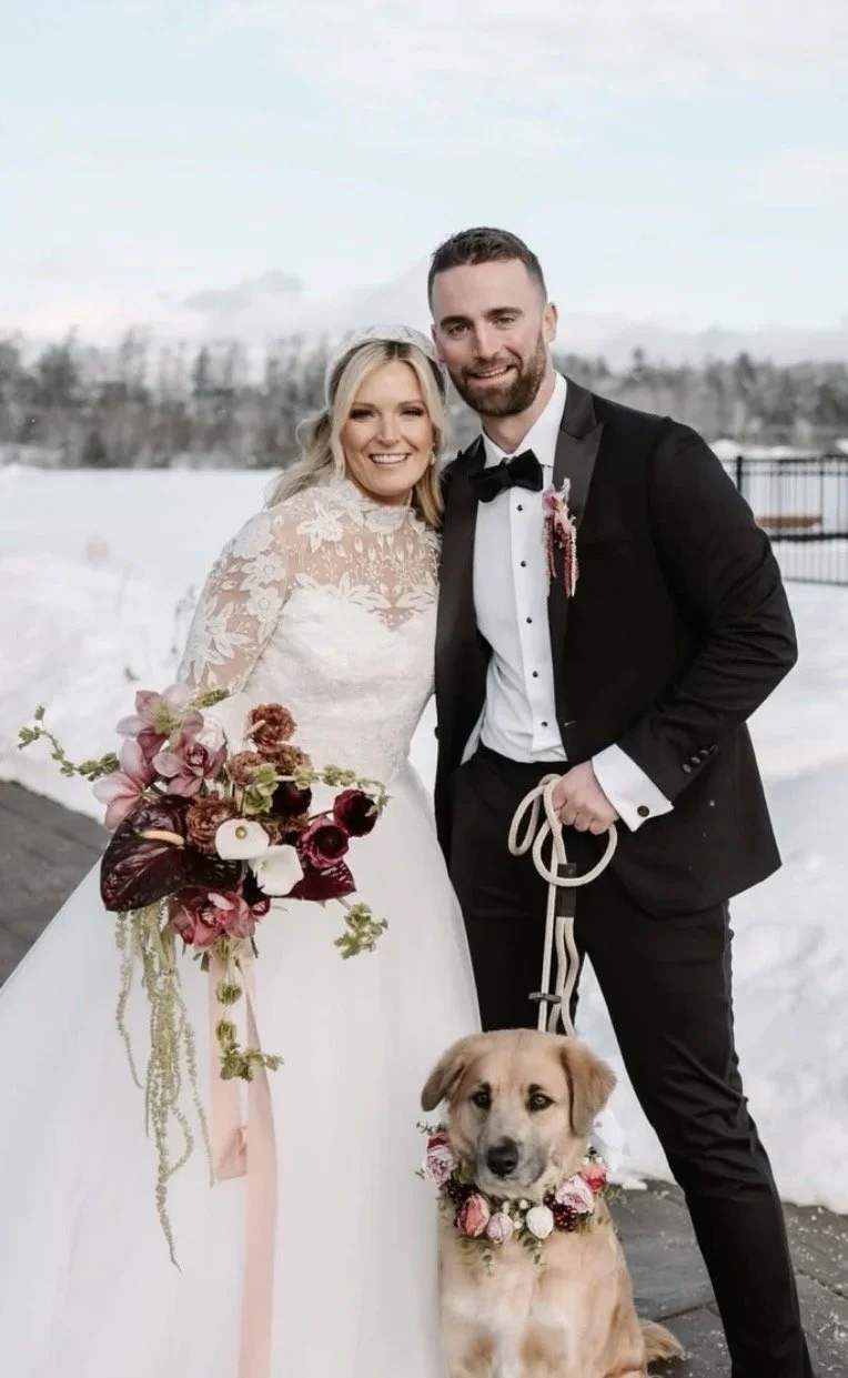A newlywed couple in wedding attire stands outdoors in a snowy landscape, smiling at the camera with their dog sitting in front of them, decorated with a floral collar. The bride holds a bouquet of flowers, and the groom is dressed in a tuxedo.