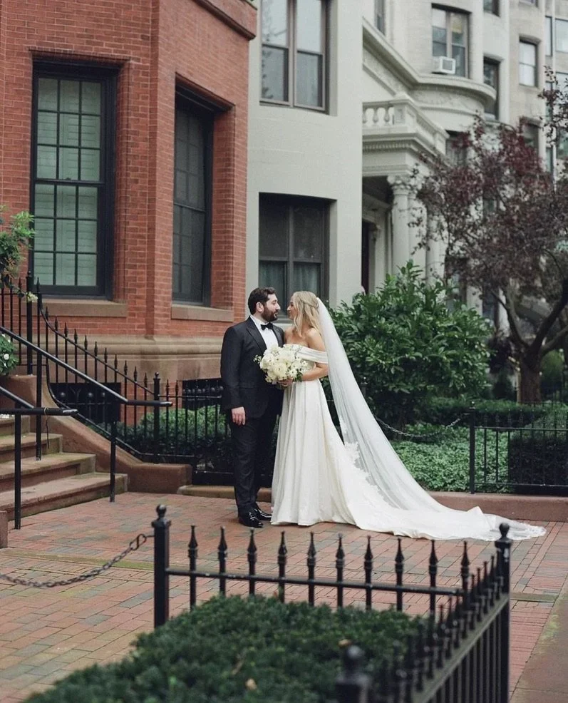 A bride and groom standing close together outdoors in front of a building with brown and white facades, surrounded by greenery, with the bride holding a bouquet of white flowers.