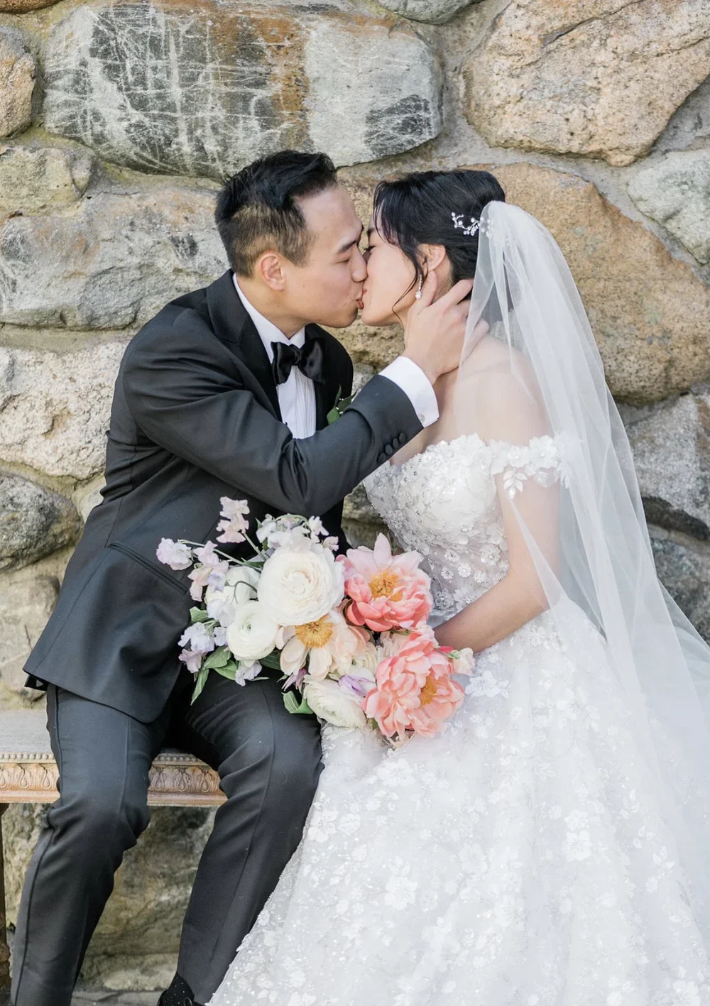 A newlywed couple sharing a romantic kiss, with the groom in a black tuxedo and the bride in a white wedding gown holding a bouquet of pink and white flowers, against a stone wall background.