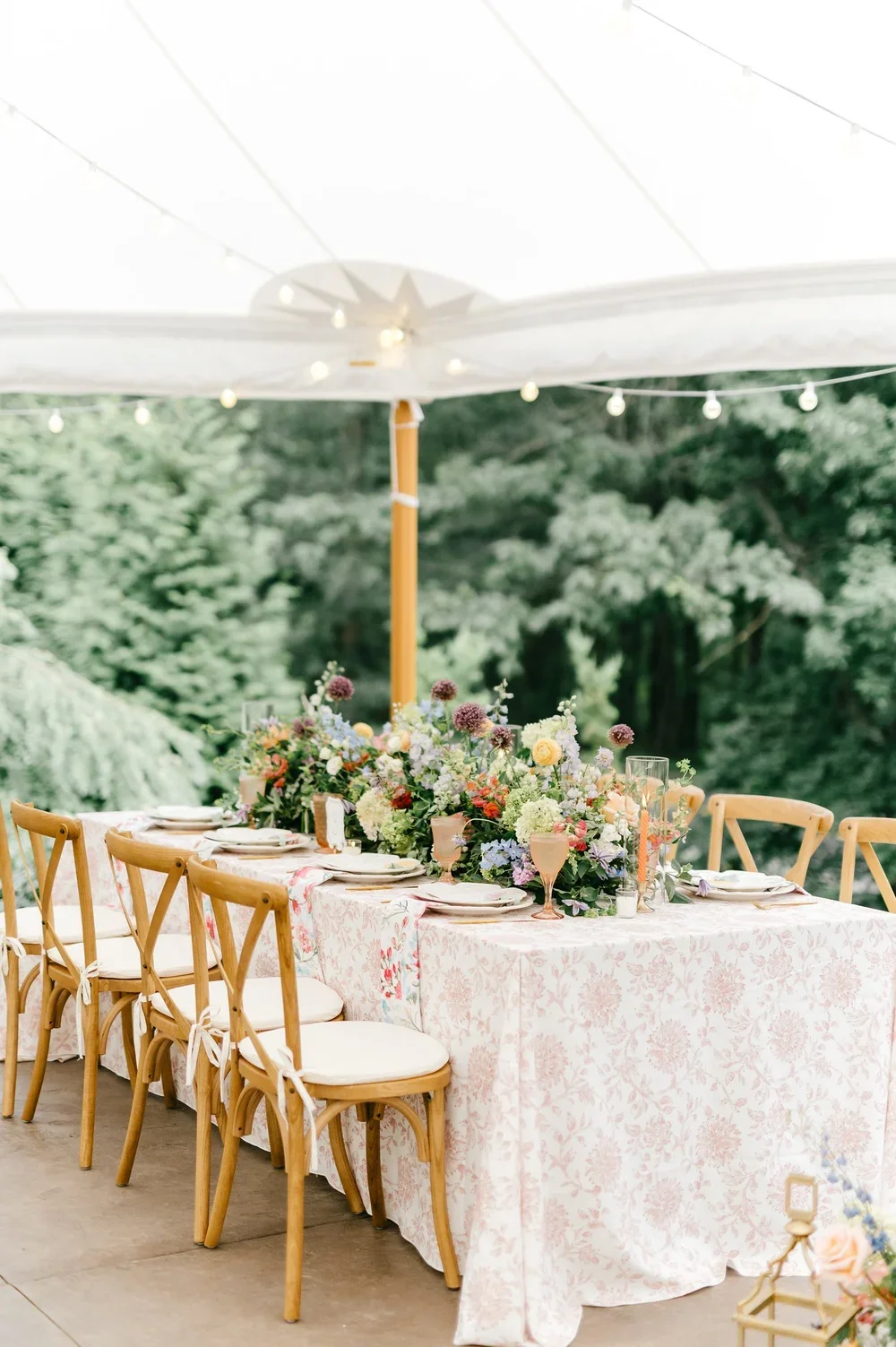 Outdoor dining setup under a white canopy with a long table decorated with a floral centerpiece, surrounded by wooden chairs with white cushions.