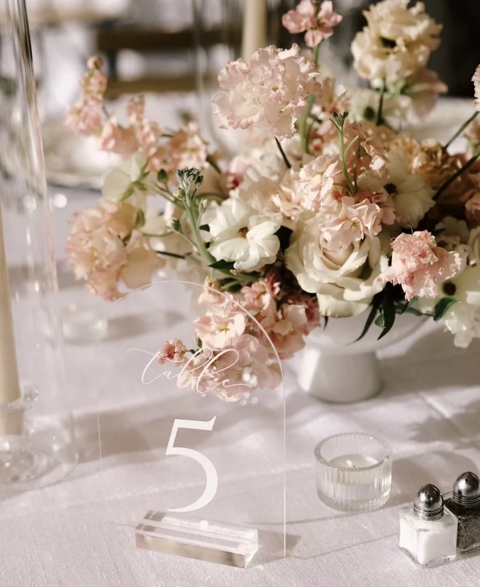 A table decorated with a floral centerpiece of pink and white flowers, a clear table number card with the number 5, and small containers of salt and pepper.