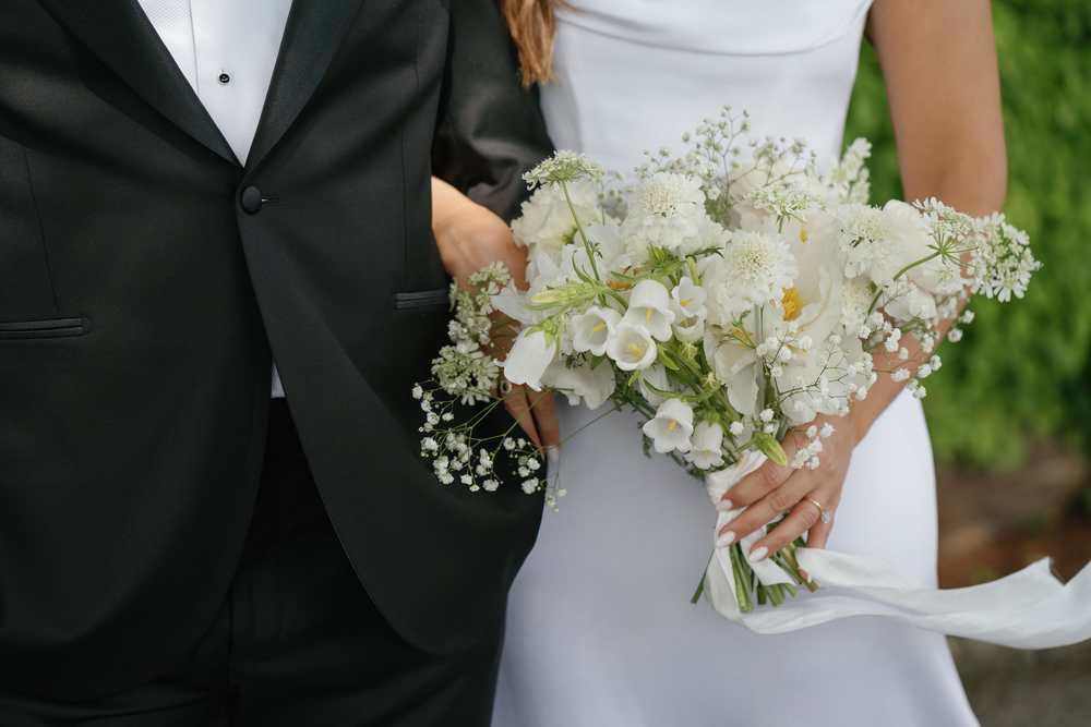 Close-up of a bride holding a bouquet of white flowers, standing next to a groom in a black tuxedo outdoors.