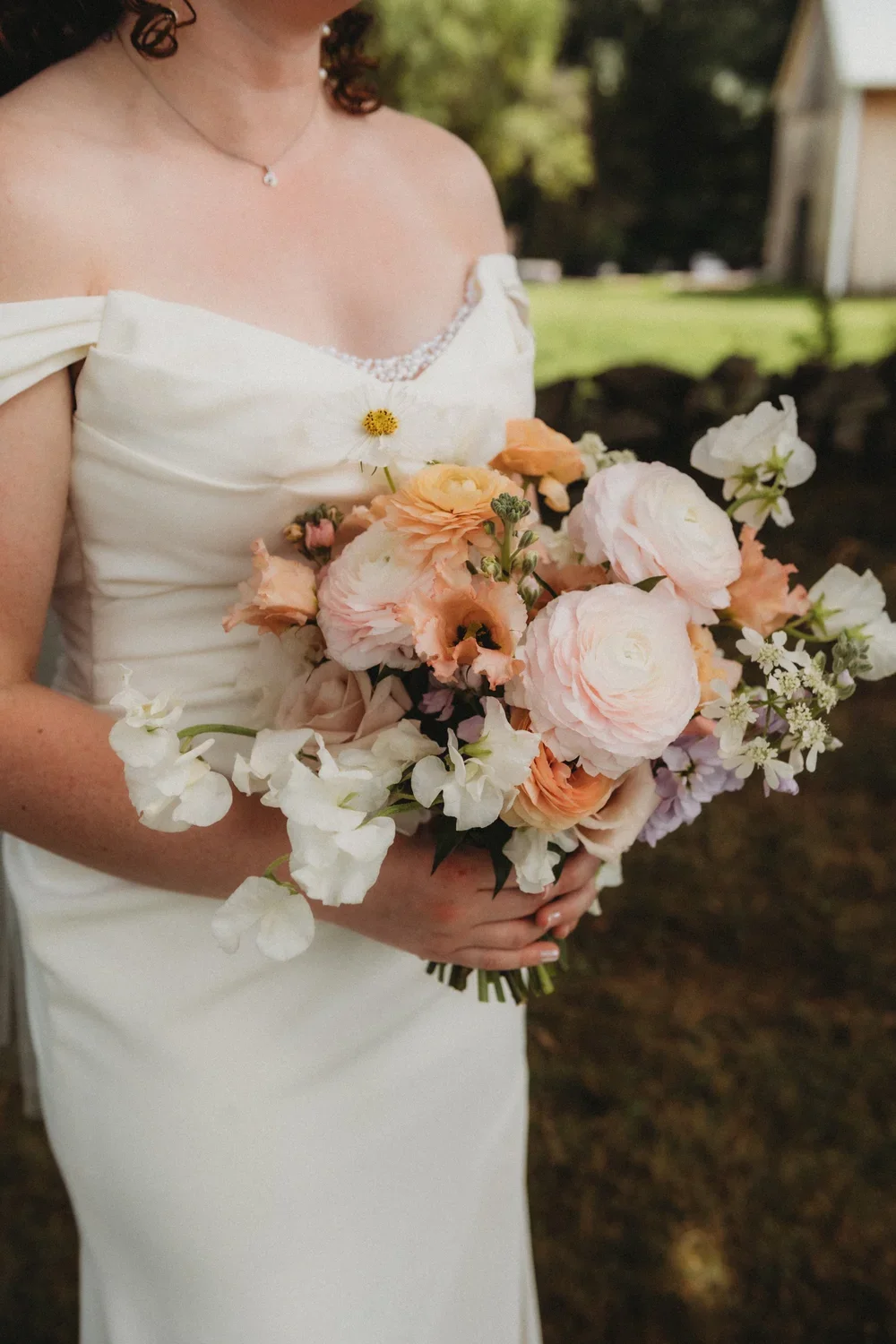 A bride in a white wedding dress holding a bouquet of peach, white, and pink flowers outdoors.