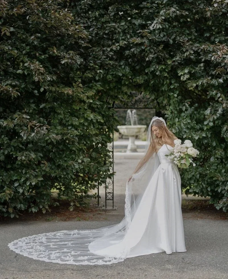 Bridal photo of a woman in a white wedding dress holding a bouquet, standing outdoors near greenery and a fountain in the background.