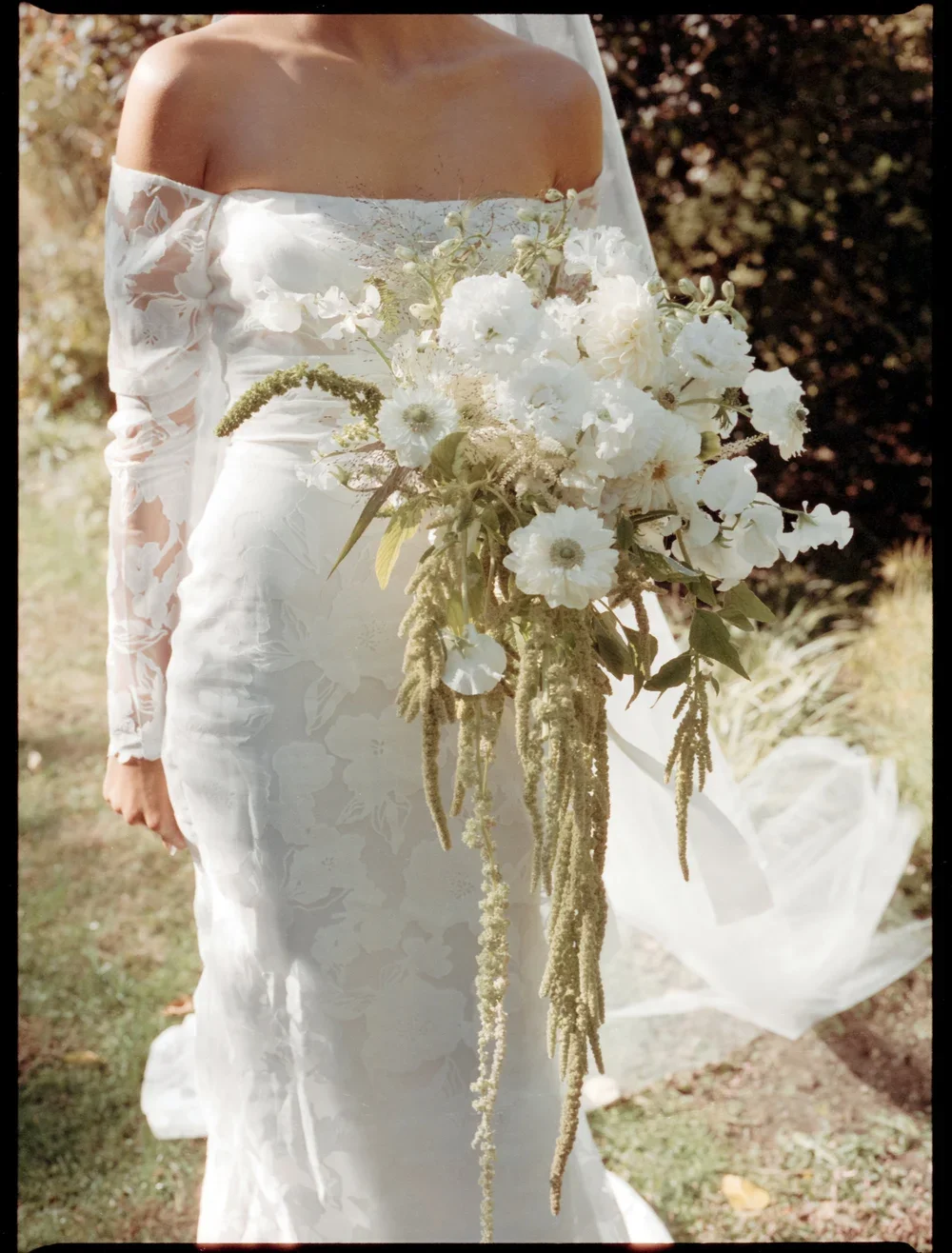 A bride in a white dress with off-shoulder lace sleeves holding a cascading bouquet of white and green flowers outdoors.