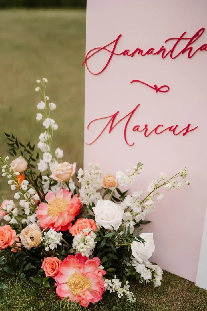 A pink wedding sign with red cursive writing that says 'Samantha & Marcus,' decorated with a floral arrangement of white, pink, and peach flowers and greenery, set outdoors on grass.