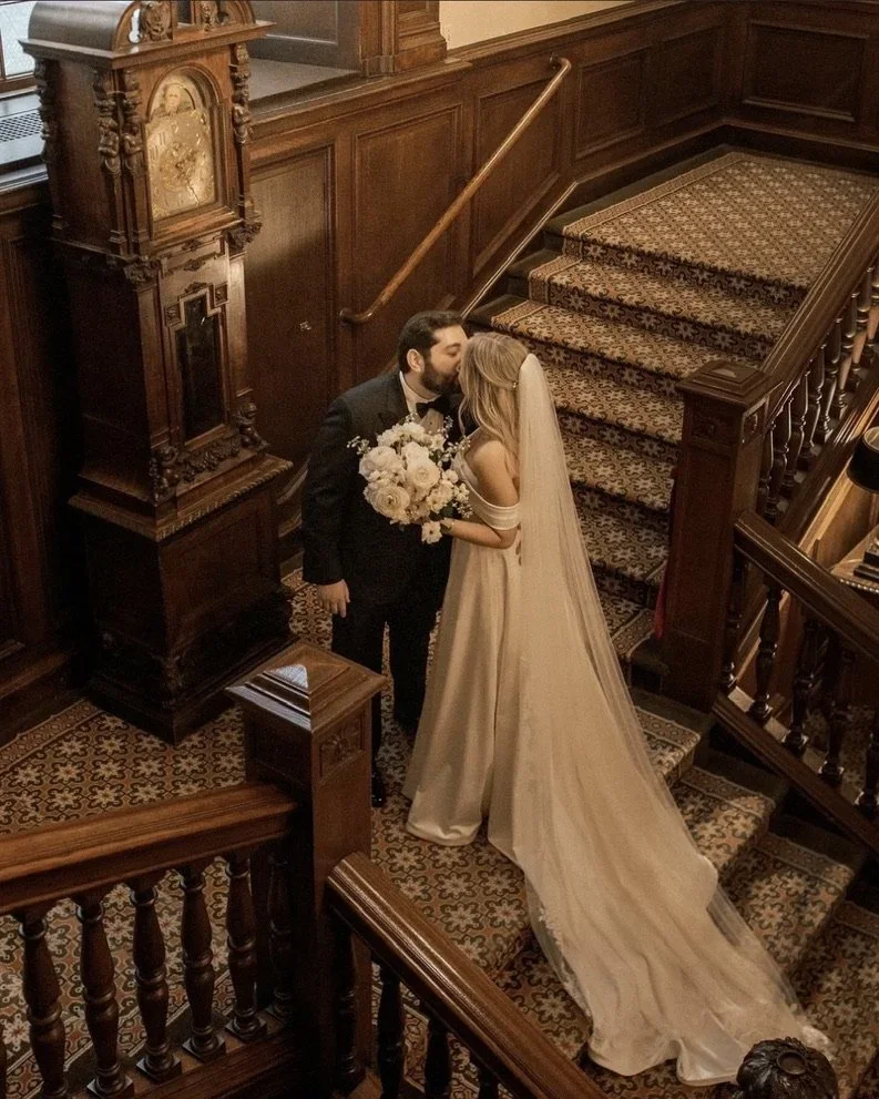 A bride and groom sharing a kiss on a staircase indoors, with ornate wooden paneling and a large floral bouquet, in a vintage or historic building setting.