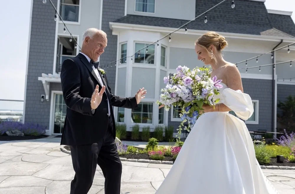 A bride in a white wedding dress holding a bouquet of flowers looks happy as she dances with an older man in a tuxedo in an outdoor setting decorated with string lights.