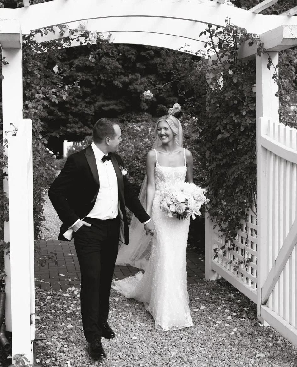 A black-and-white photo of a bride and groom walking through a garden archway. The bride is smiling and holding a bouquet, wearing a lace wedding dress, while the groom, dressed in a tuxedo with a bow tie, is looking at her and holding her hand.