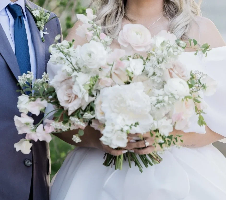 Bride holding a bouquet of white and light pink flowers, with groom partially visible on her left.
