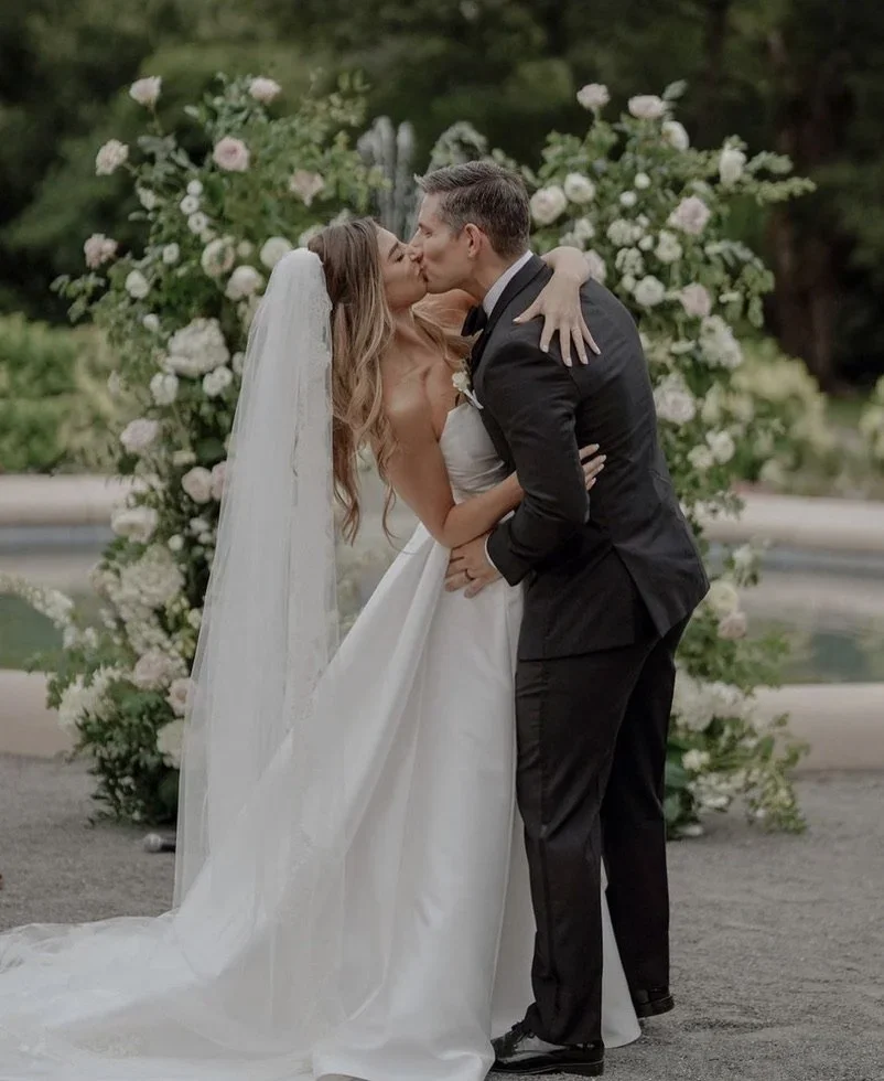 A bride and groom share a kiss during their wedding ceremony, standing in front of a floral arch decorated with white and pink flowers, outdoors.