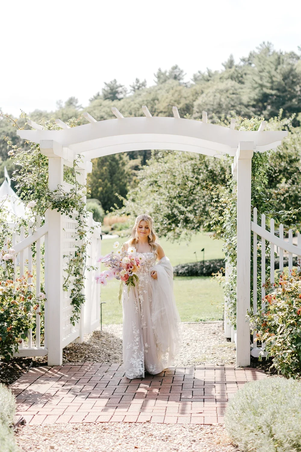 A bride holding a bouquet of pink and purple flowers walks through a white garden archway decorated with climbing plants, in a lush outdoor setting on a sunny day.