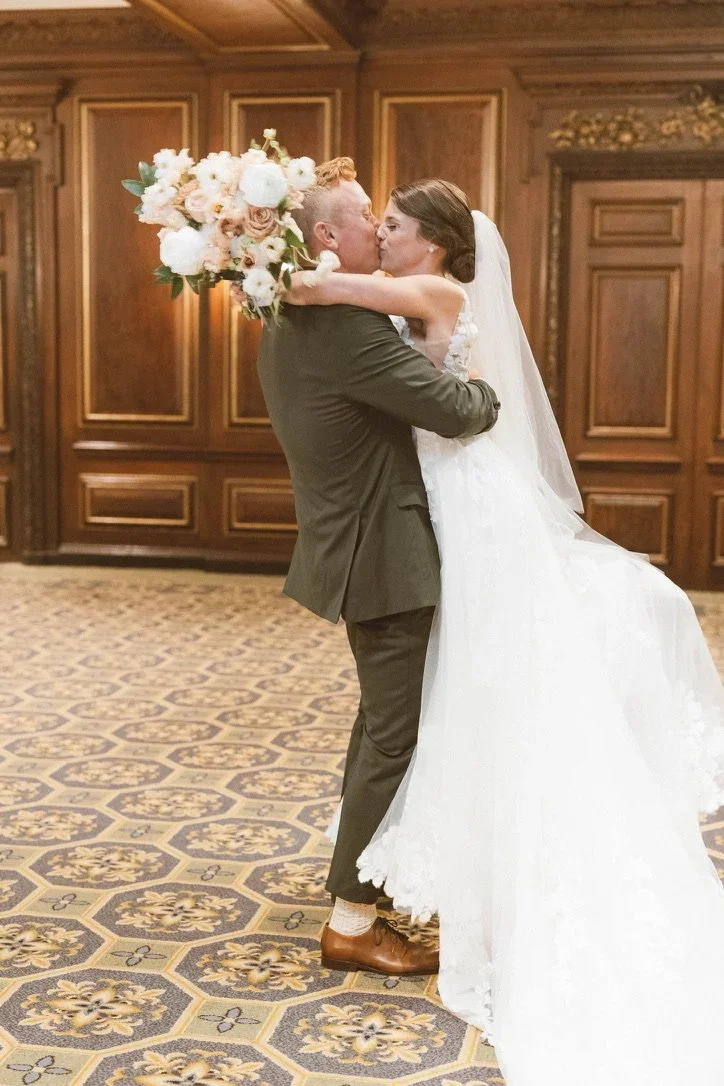A bride and groom share a kiss at their wedding ceremony, with the bride holding a large bouquet of flowers, in a wood-paneled room with ornate decor.