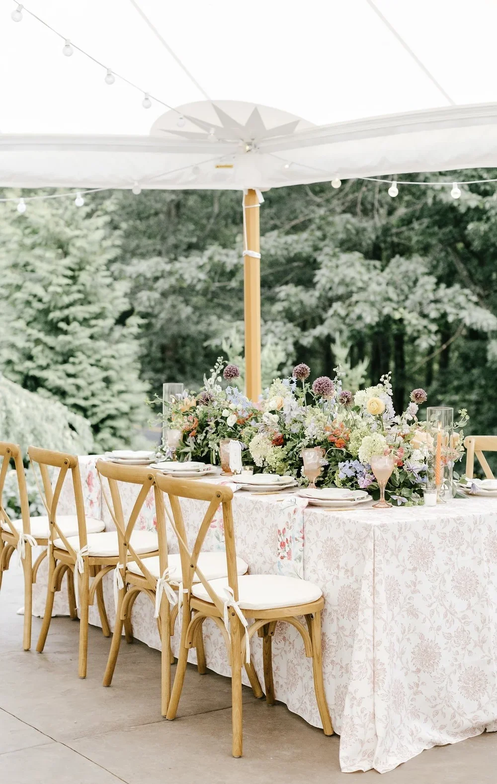An outdoor dining setup with a long table decorated with flowers and candles, under a large white umbrella, surrounded by wooden chairs with white cushions, in a lush green environment.