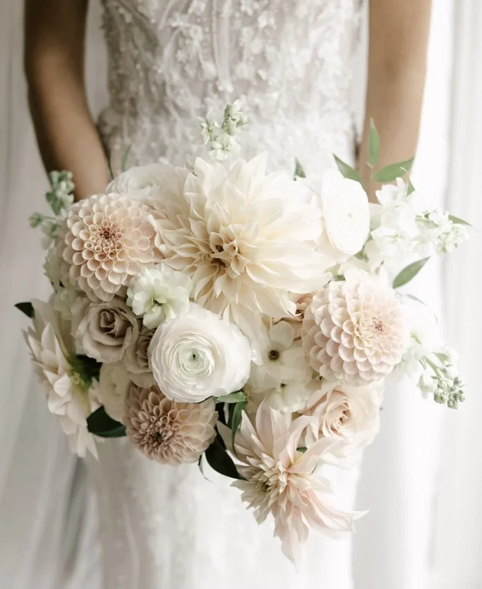 A bride holding a bouquet of white and blush-colored flowers, including roses, dahlias, and ranunculus, in front of a white backdrop.
