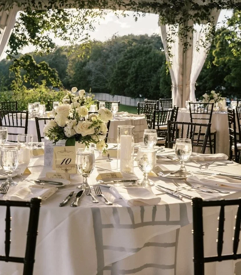 Elegant outdoor wedding reception setup with round tables covered in white tablecloths, decorated with floral centerpieces, candles, glassware, and silverware. Black chairs surround the tables under a draped tent with greenery outside.