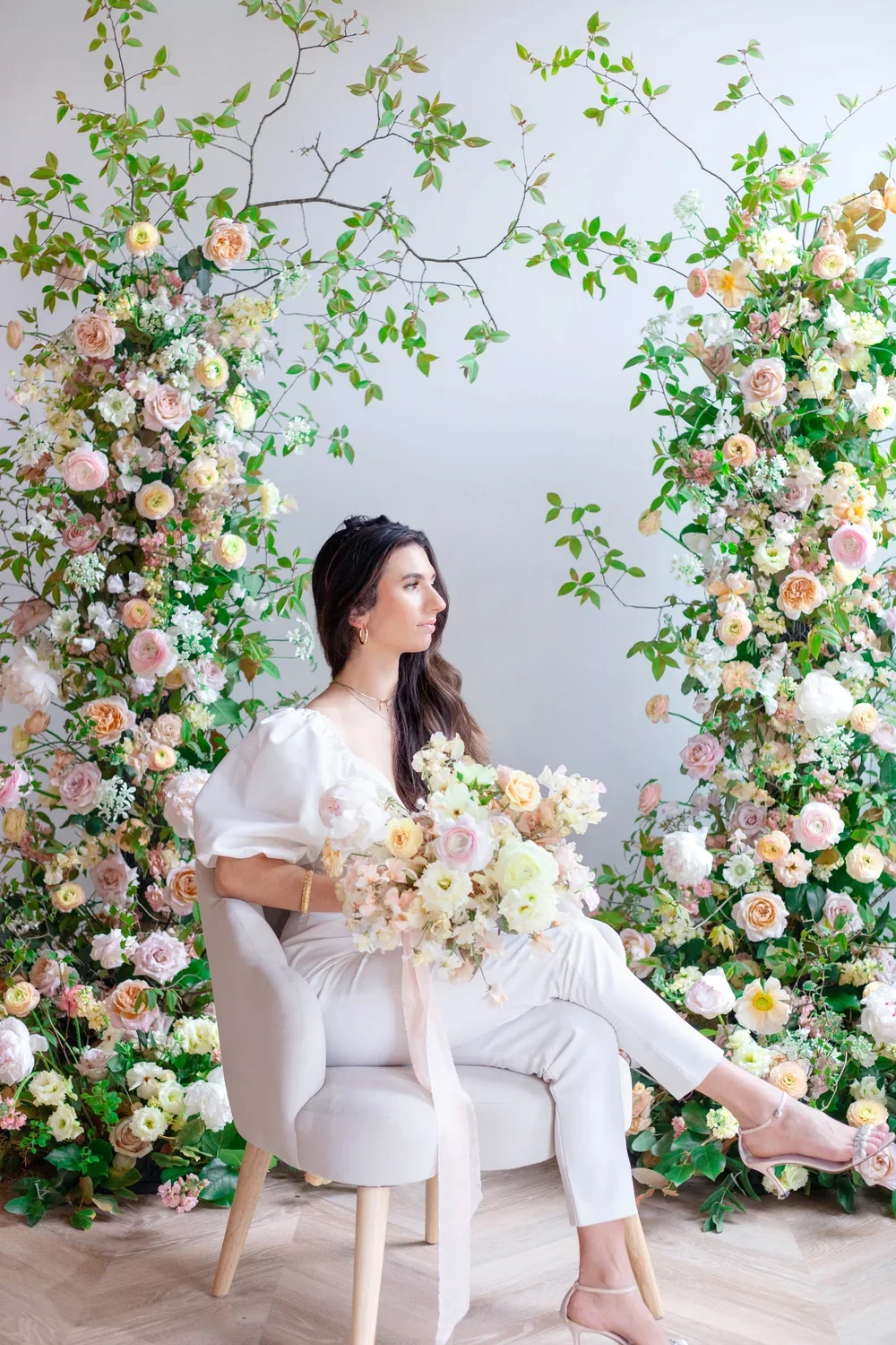 A woman with long dark hair, wearing white clothes and beige heels, is sitting on a small white chair surrounded by a floral backdrop of pink, white, and peach roses and greenery.