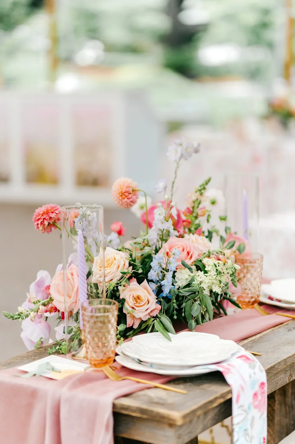 A floral centerpiece on a wooden table with pastel-colored roses, eucalyptus, and other flowers, surrounded by pink glass candle holders, set for a celebration or event in a bright, airy room.