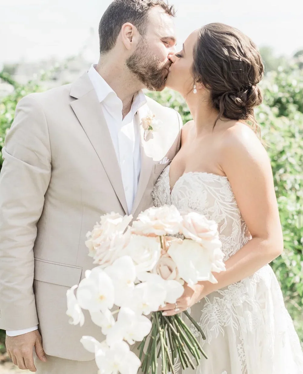 A newlywed couple sharing a kiss outdoors with a bouquet of light-colored flowers.