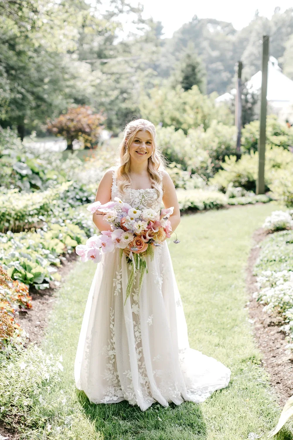 A smiling bride in a white lace wedding dress holding a colorful bouquet of flowers outdoors in a garden.
