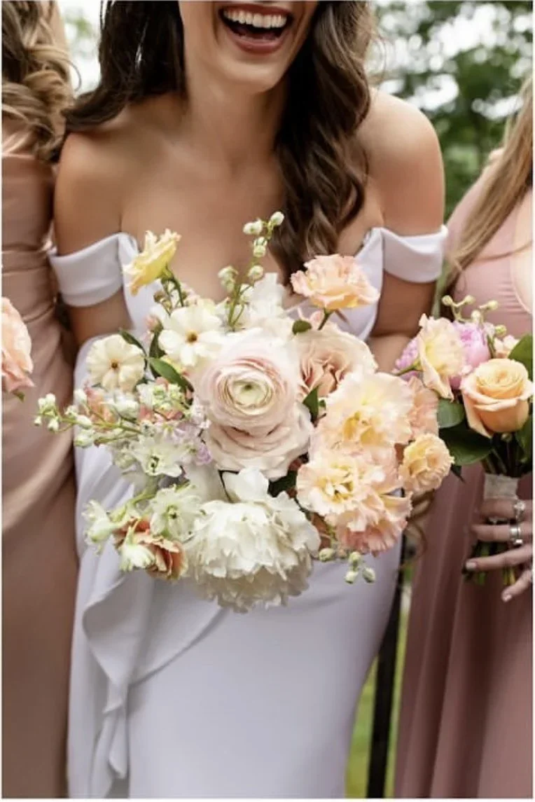 A woman in a white dress holding a bouquet of pink and white flowers, smiling with visible teeth, surrounded by other women in pastel dresses.