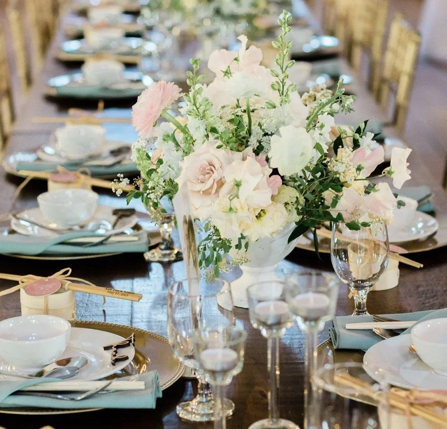 A formal dining table decorated with a central flower arrangement of pink and white roses, lilies, and greenery, surrounded by white plates, silverware, aquamarine napkins, wine glasses, and small candles in glass holders.