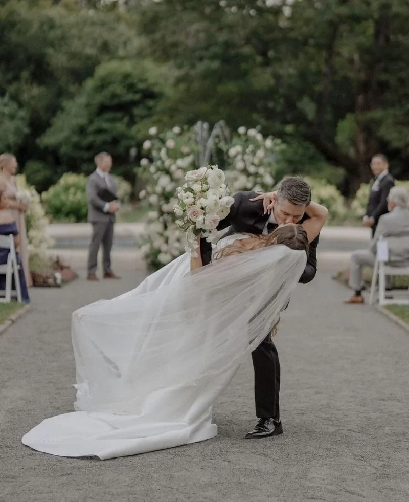 Groom dips bride during wedding ceremony outdoors, with guests seated and standing nearby, greenery in the background, and a decorative floral arrangement