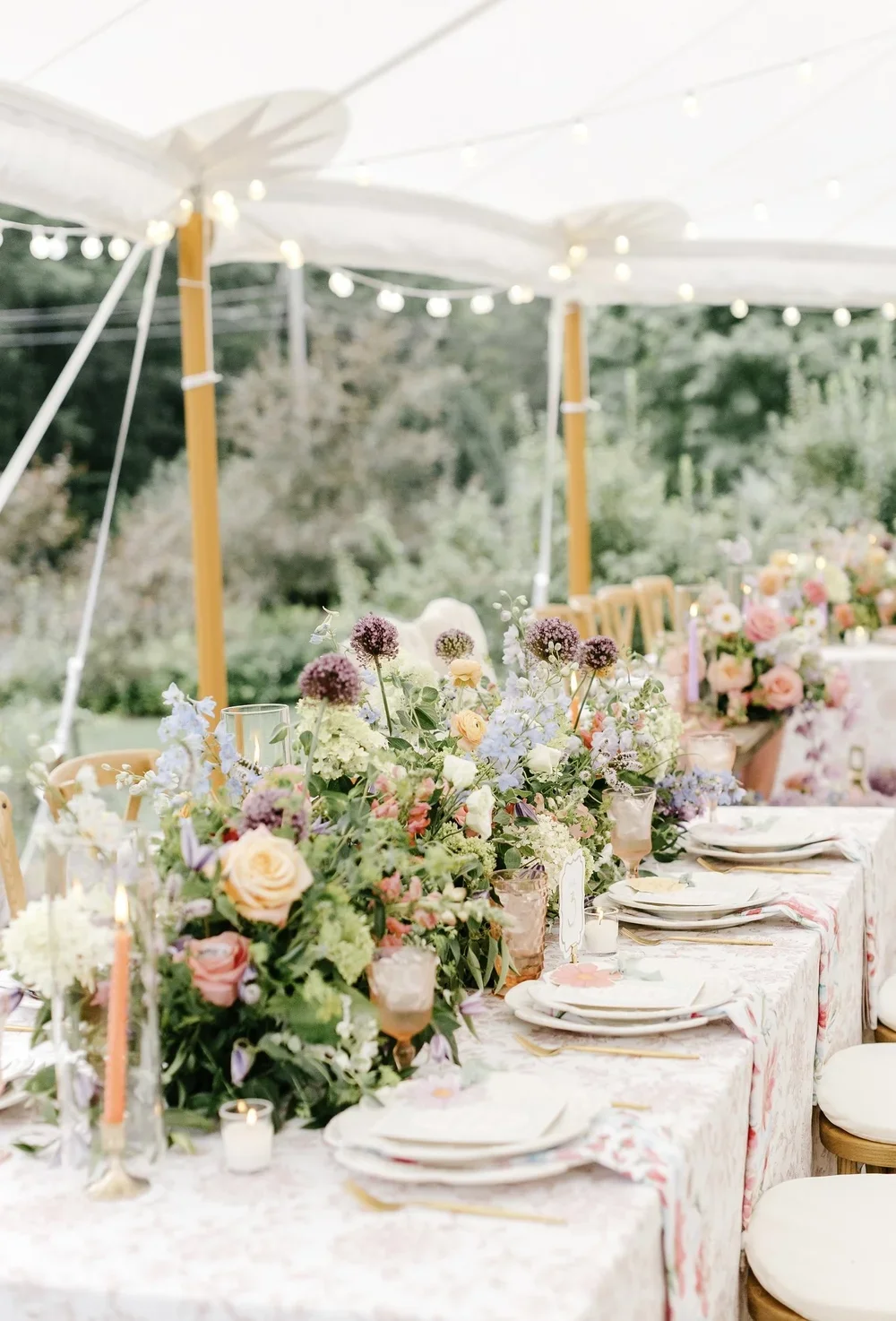 Decorated outdoor dining table with floral arrangements, candles, and tableware, under a canopy with string lights at sunset.