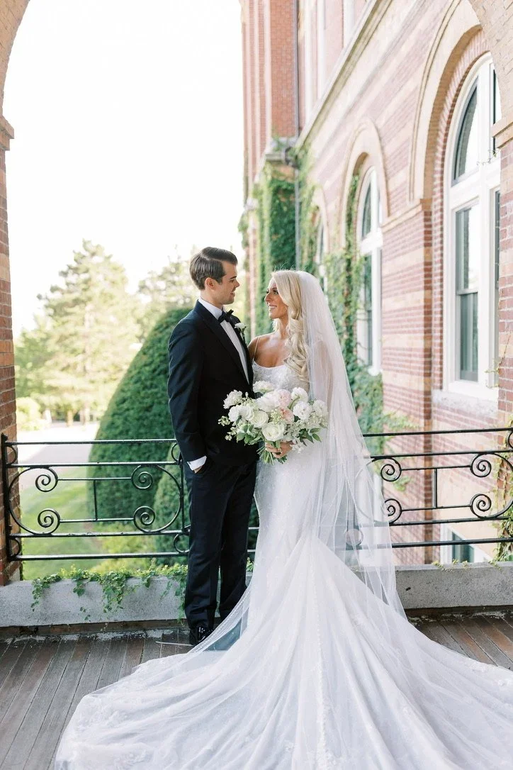 Bride and groom standing on a balcony, gazing at each other. The bride is holding a bouquet of white and pink flowers, wearing a white wedding dress with a long veil. The groom is dressed in a black tuxedo with a bow tie. The background features a br