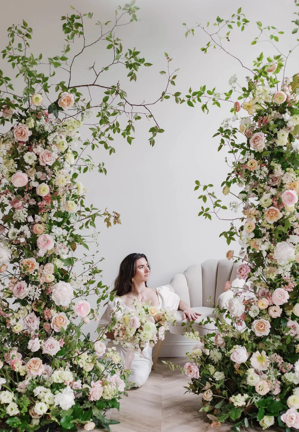 A woman dressed in white posing on a beige sofa, surrounded by an arch of pink and white flowers and green foliage against a plain white wall.
