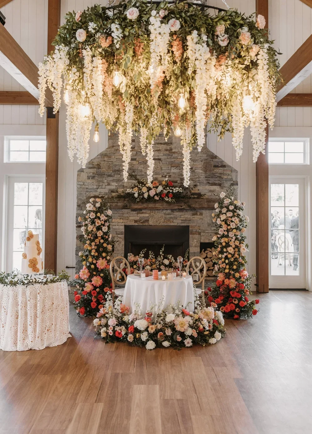 Elegant wedding reception area with a stone fireplace decorated with flowers, a round cake with golden accents, and a floral chandelier hanging from the ceiling.