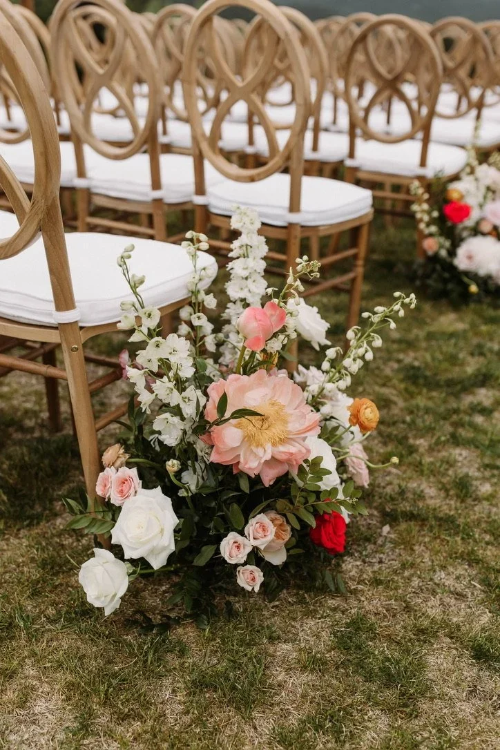 A floral arrangement with pink, white, and red roses and white flowers on the ground in front of wooden chairs with white cushions, set outdoors on grass.
