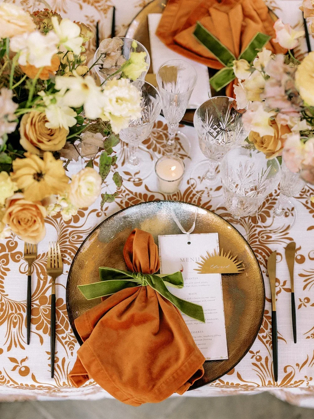 Elegant table setting with floral centerpieces, glassware, gold cutlery, a golden charger, a folded orange napkin tied with a green ribbon, a printed menu, and a lit candle.