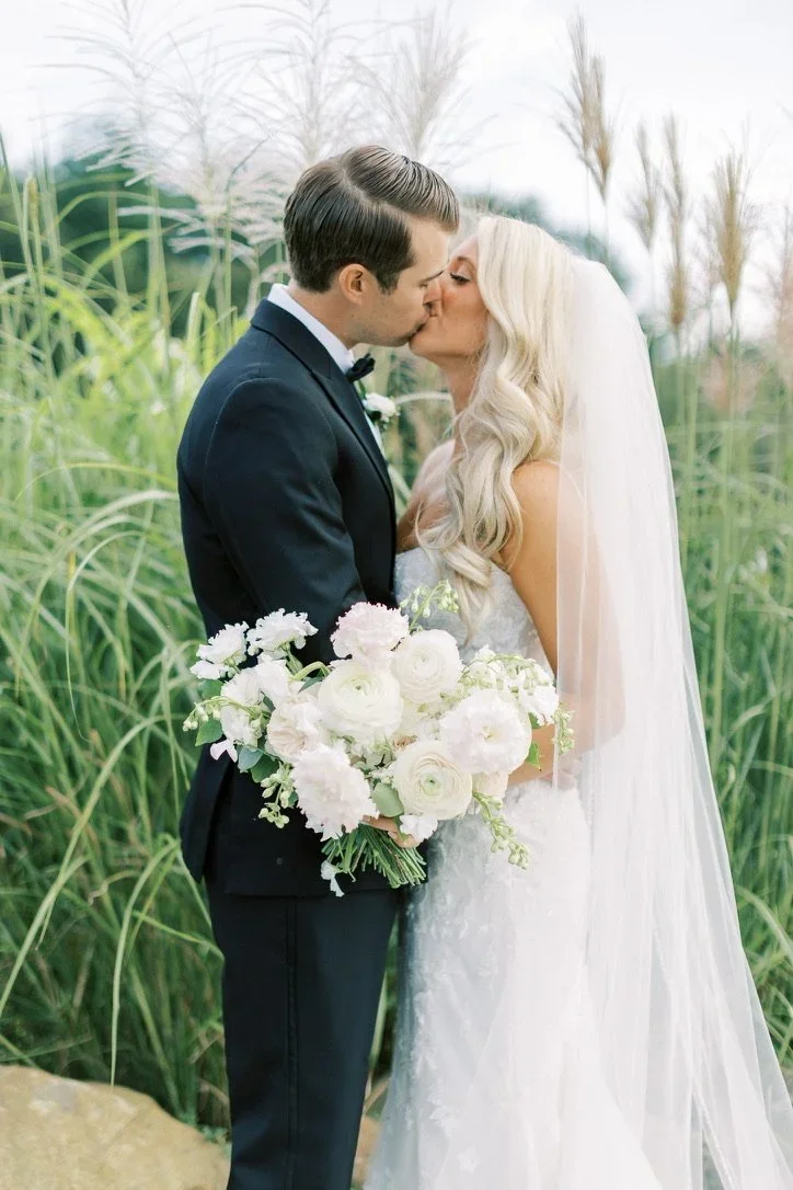 Bride and groom sharing a kiss in a natural outdoor setting, with the bride holding a bouquet of white flowers.