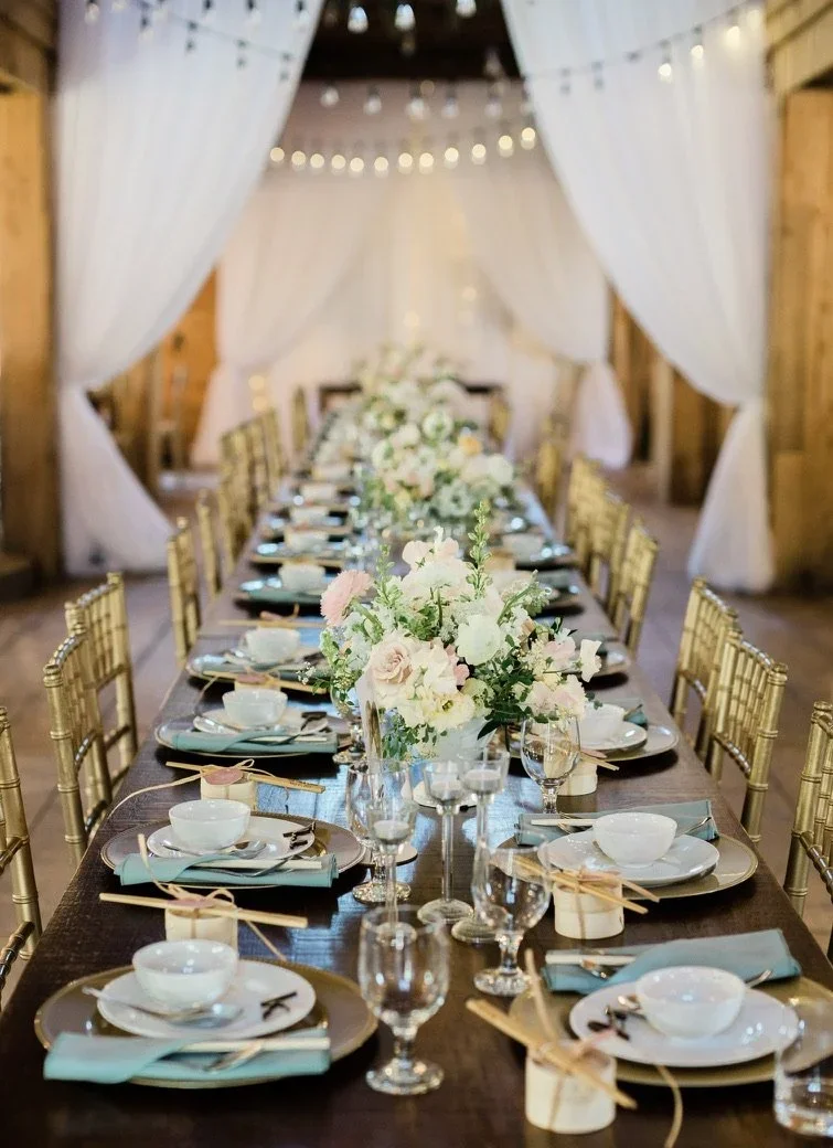 A long banquet table set for a celebration with white and pastel floral centerpieces, gold chairs, and elegant tableware, in a rustic room with string lights and draped white curtains.