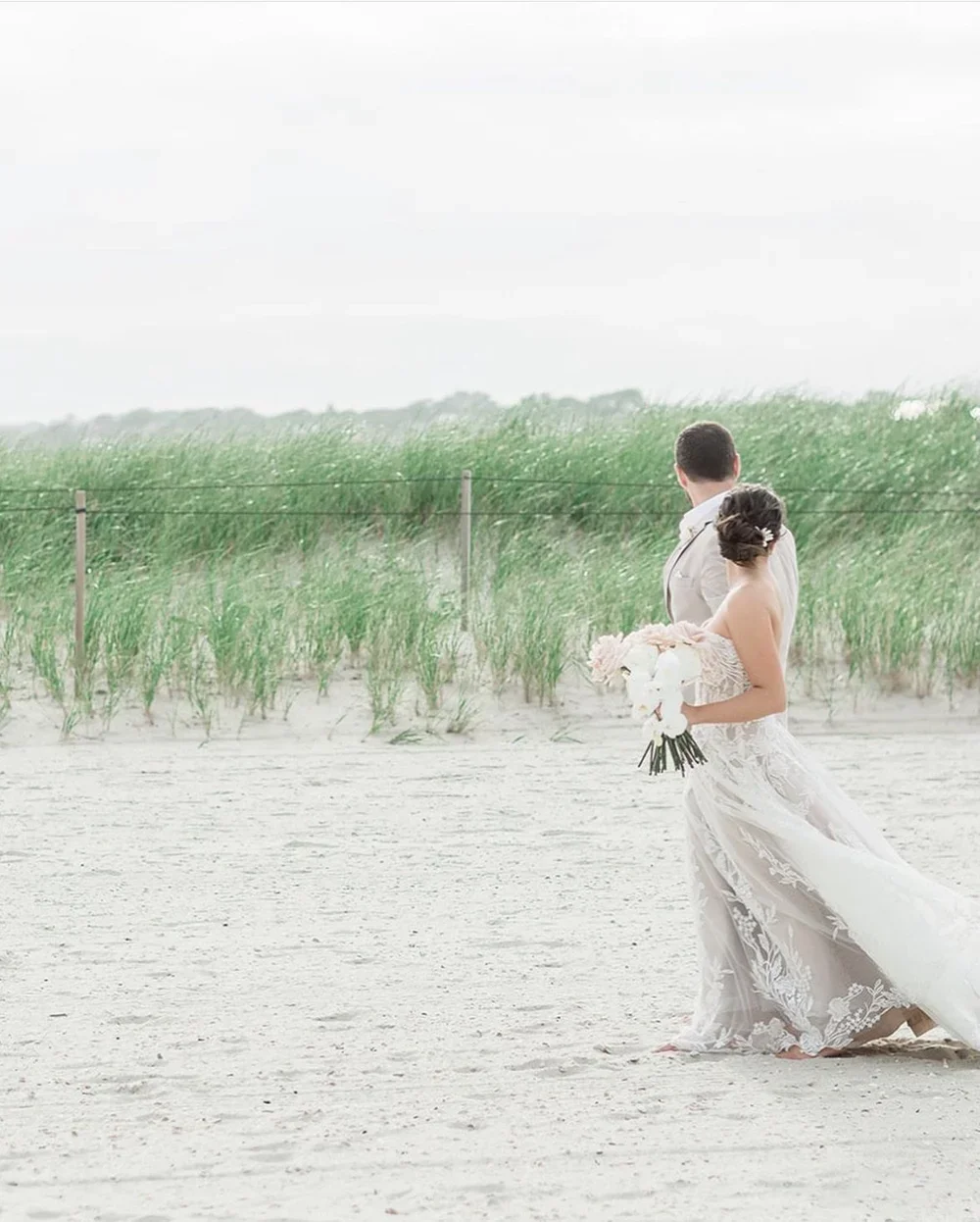 A bride and groom standing on a sandy beach, with the bride holding a bouquet of white flowers, during a wedding photoshoot.