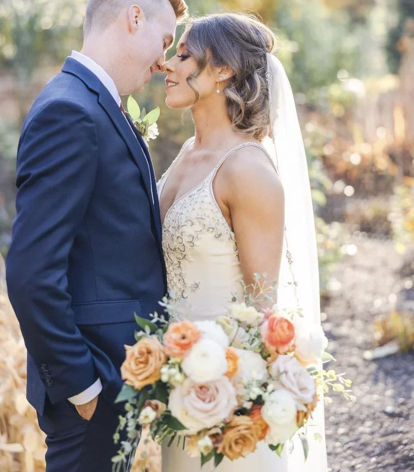 A bride and groom face close with foreheads touching, outdoors on their wedding day, the bride holding a bouquet of orange, peach, and white flowers, both smiling softly.