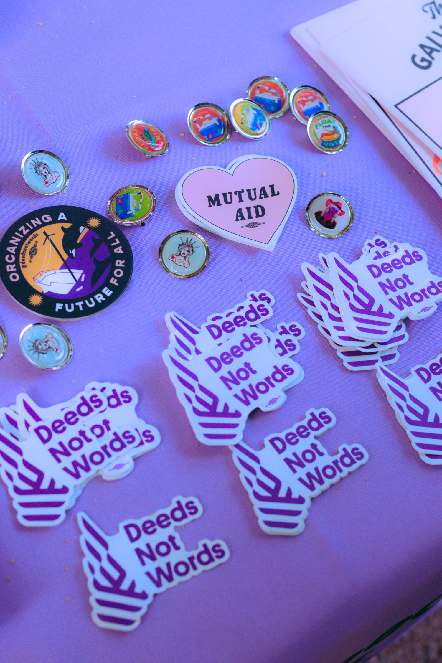 Deeds Not Words merch and stickers on a purple cloth on a table