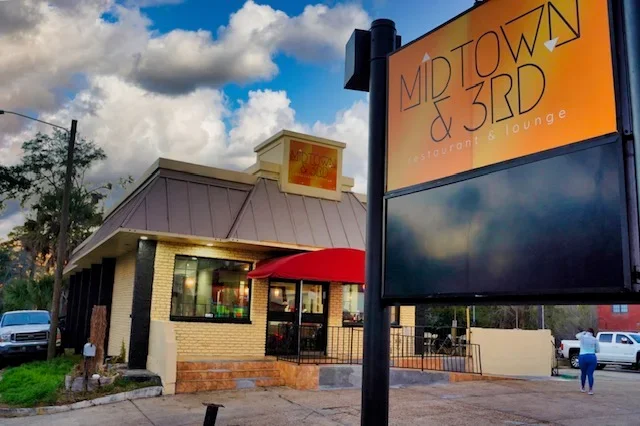 Outdoor view of Midtown & 3rd restaurant and lounge with a large illuminated sign and a building with a red awning, brick walls, and a small patio area.