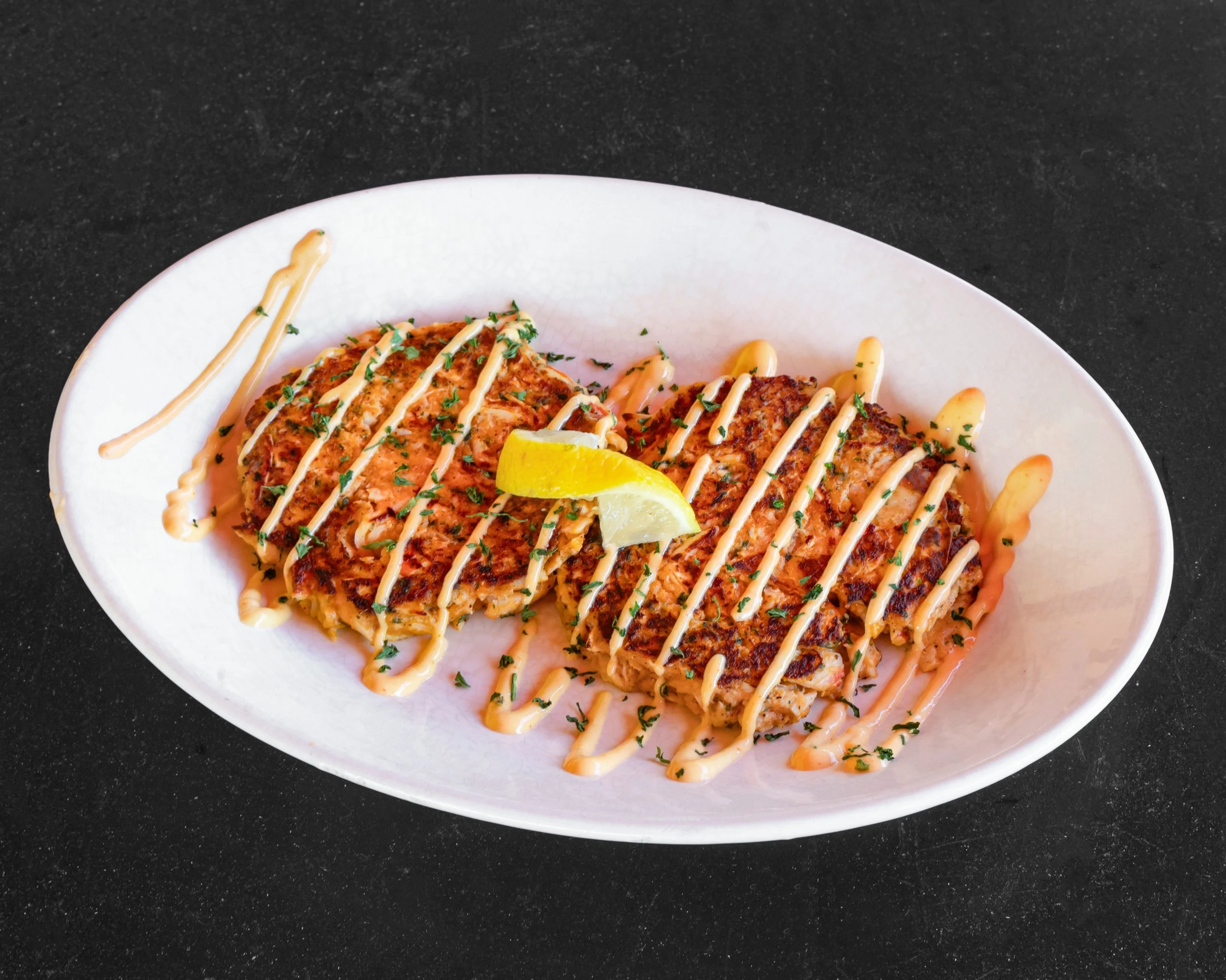 Two grilled fish fillets garnished with chopped herbs and drizzled with a creamy sauce, topped with a lemon wedge, on a white oval plate.
