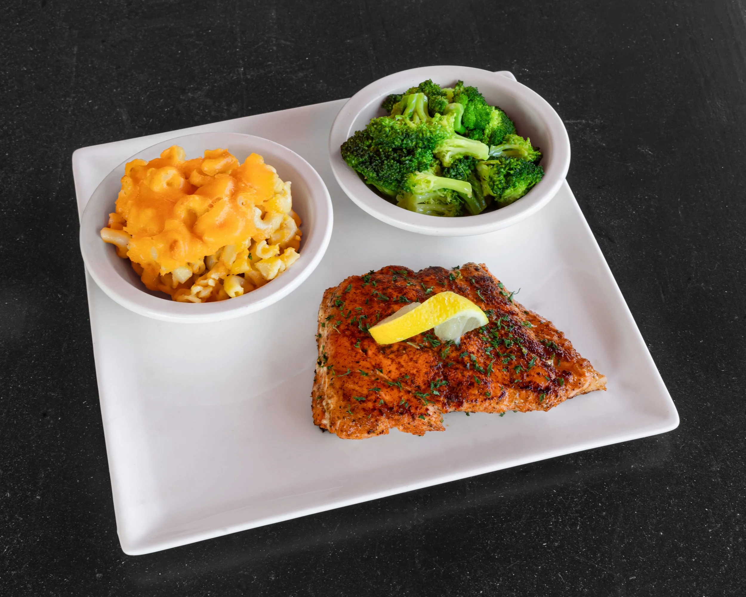 Plate with grilled fish fillet garnished with lemon; side of macaroni and cheese, and broccoli in small bowls.
