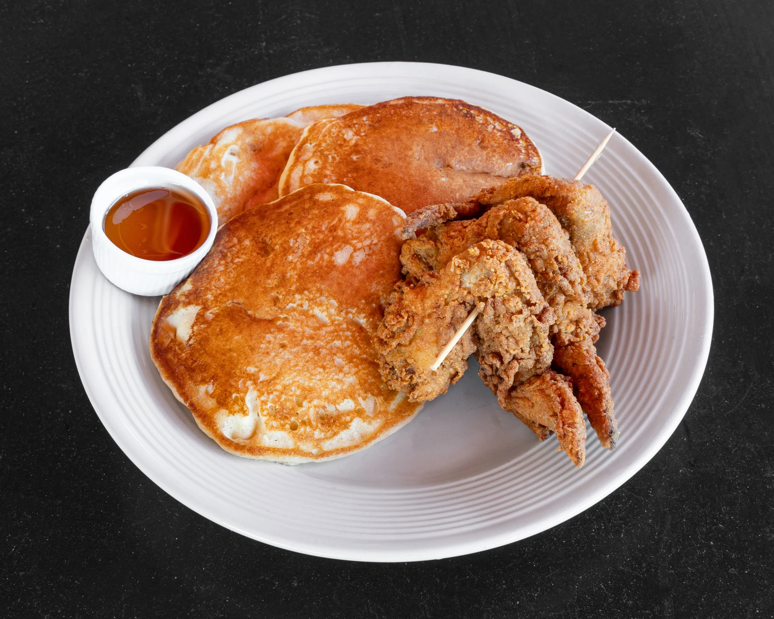 A plate of breakfast food with two fried eggs, a few pancakes, and two pieces of fried chicken, served with a small cup of syrup.