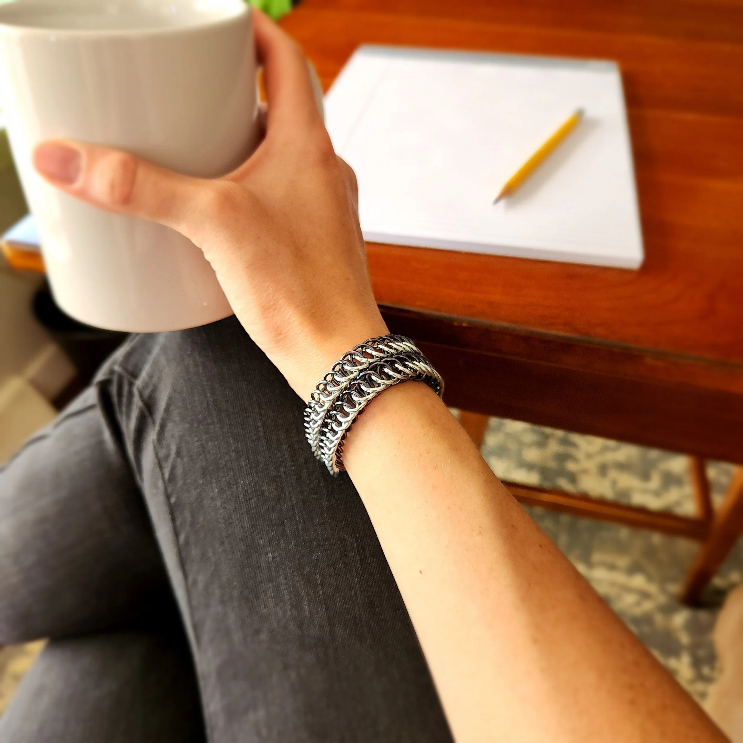 A person's arm resting on their knee, holding a white mug, with a silver bracelet on their wrist. There is a notebook with a yellow pen on a wooden table in the background.