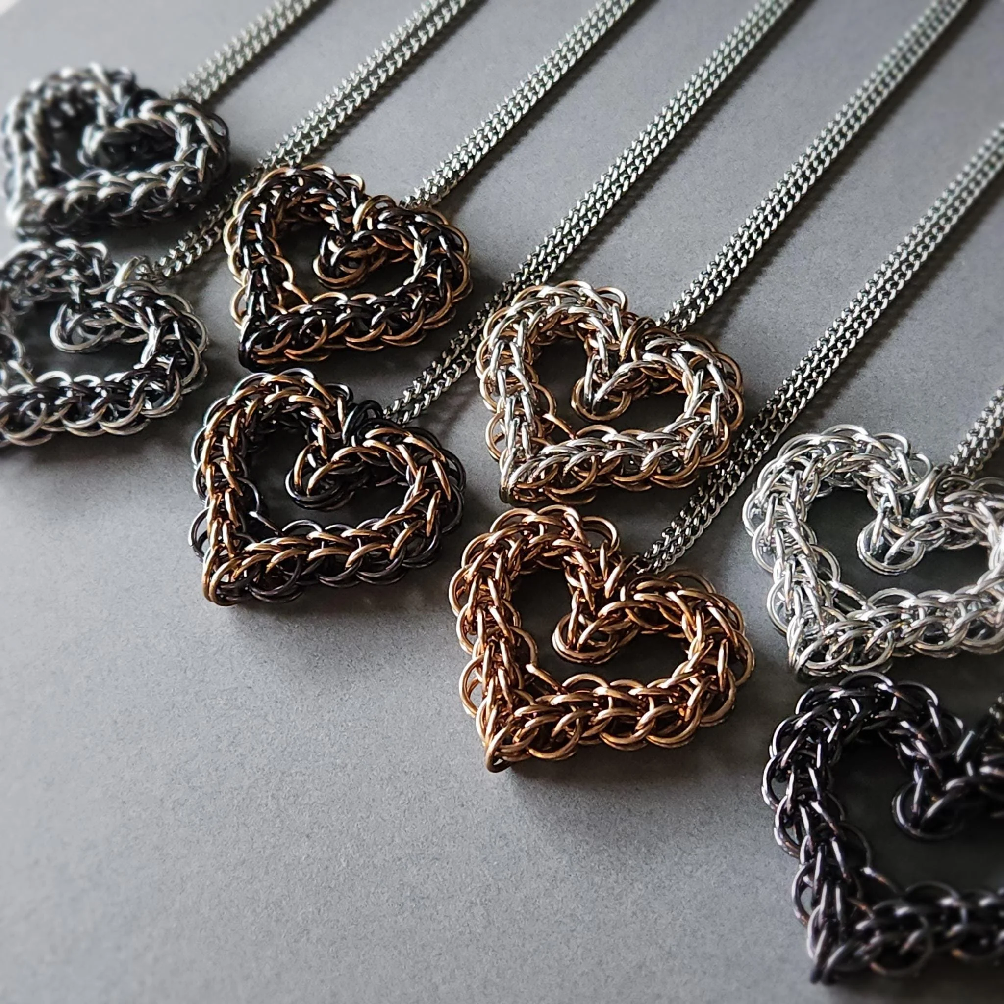 Collection of chain necklaces with heart-shaped pendants in silver, black, and rose gold tones on a gray background.
